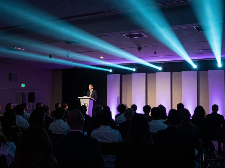 Wide-angle view of a corporate event ballroom mid-program, a speaker at a podium under tight stage lighting, audience in sharp silhouette, teal and purple event lighting raking across the space, journalistic framing from the back third of the room