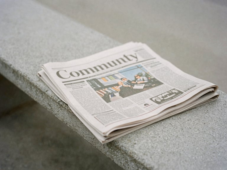 Close-up of a printed community newspaper on a concrete bench, soft overcast daylight, muted desaturated tones, quiet editorial mood.