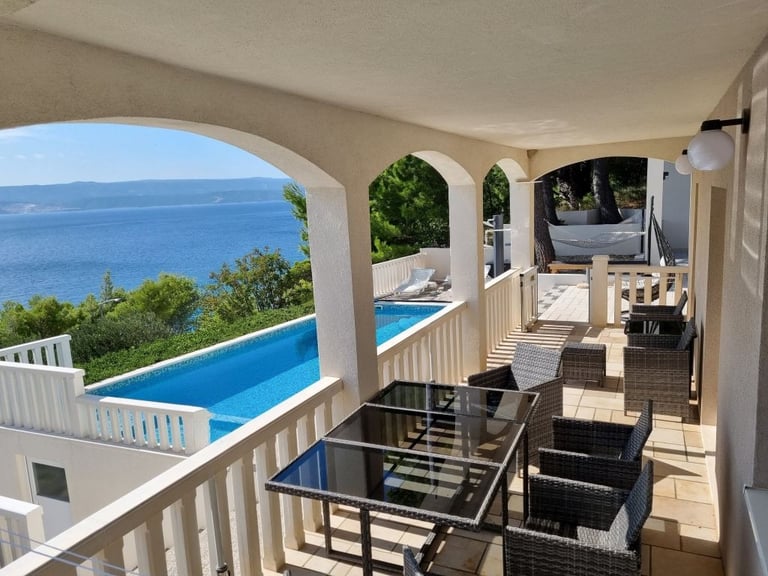 The photo shows an apartment balcony with a dining set and a view of the pool and the sea.