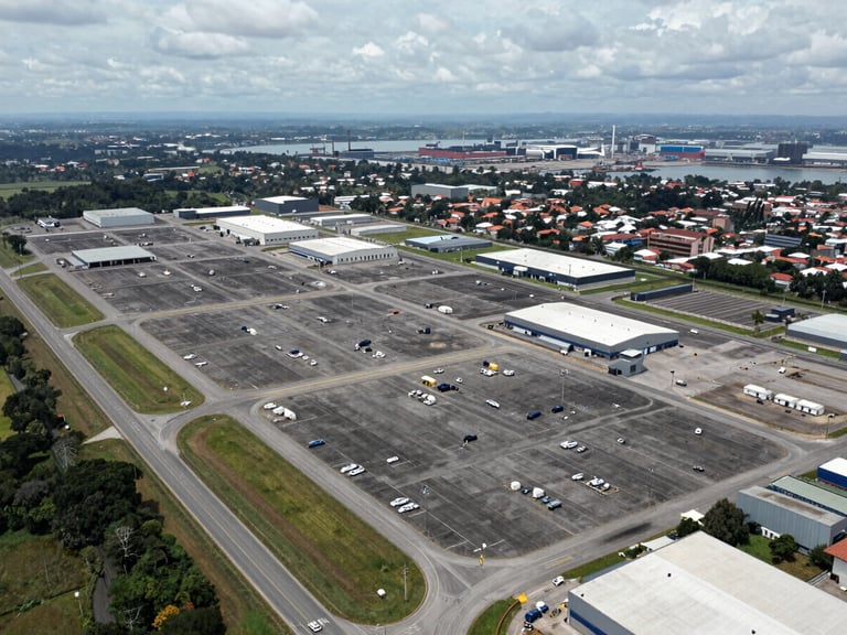 Elevated wide-angle aerial photograph of a 200-hectare industrial logistics platform in Apartadó, Antioquia, Colombia, shot from above looking south-east, paved internal roads forming a grid pattern across the site, warehouse pads and utility corridors clearly delineated, Route 62 highway visible along the left edge, port infrastructure on the far horizon, overcast natural daylight for maximum detail clarity, no people, no vehicles