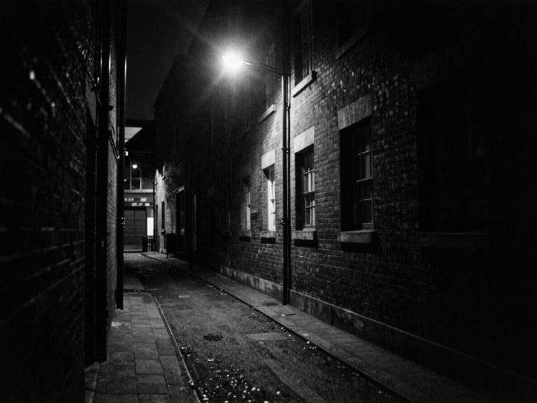 Gritty black and white 35mm film shot of a dark London alleyway at night under a single harsh street lamp, deep shadows, high contrast, heavy film grain.