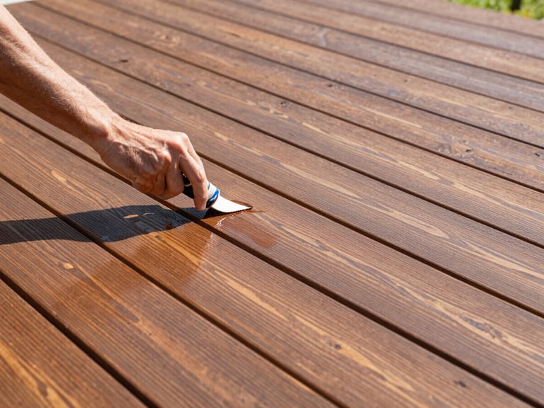 Hand applying a protective stain to a wooden deck surface, showing rich color and smooth finish, crisp outdoor photography, bright morning daylight, natural colors, sharp focus, architectural framing, 35mm lens