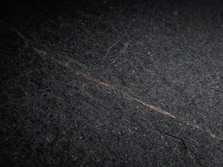 Tight macro shot of a dark, textured graphite stone surface with subtle bronze veins, dramatic side-lighting, high contrast, architectural, 35mm