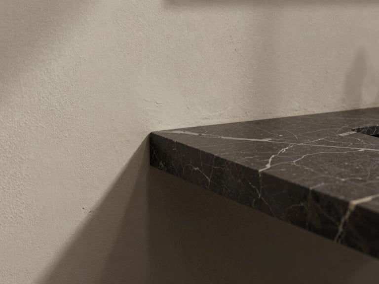 Close-up of luxury material transition, dark marble countertop meeting textured plaster wall, soft directional daylight, architectural lines, quiet luxury aesthetic.