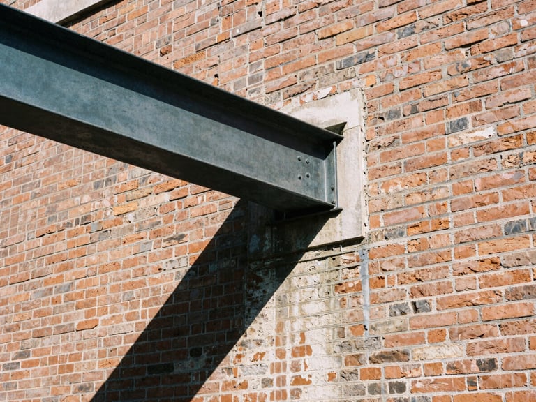 Architectural film photography, close-up details of sandblasted red brickwork meeting a massive dark steel beam, sharp natural daylight, high-contrast shadows.