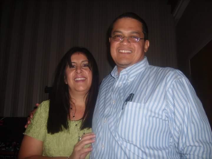 A smiling couple standing together at an indoor event, featuring a man in a blue striped shirt.