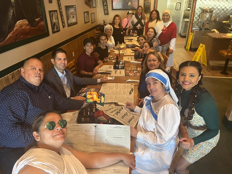 A large group of smiling friends wearing casual clothes and costumes dine together at a restaurant table.