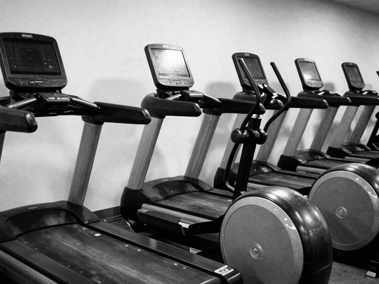 Empty cardio row with treadmills and elliptical machines, black and white, high contrast, reflections on screens, clean and spacious feel, harsh side lighting.