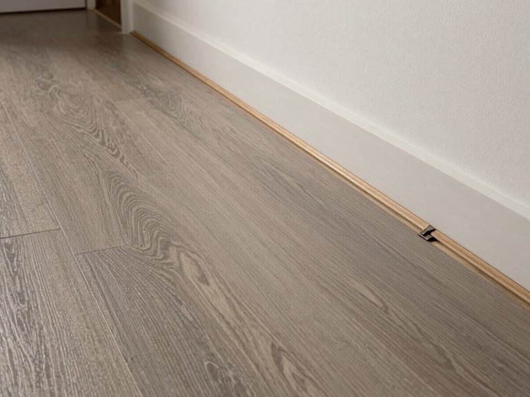 Low-angle raking shot of luxury vinyl plank flooring in a hallway, moisture barrier visible at the edge where installation begins, soft overhead light showing plank texture and locking seam, no people