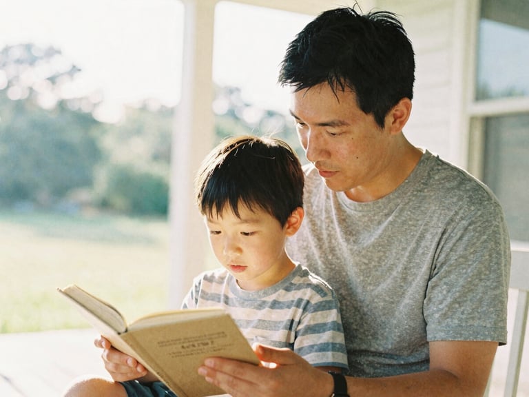 Warm editorial photography of a father and young son reading together on a sunlit porch, soft morning daylight, shallow depth of field, 35mm film style.