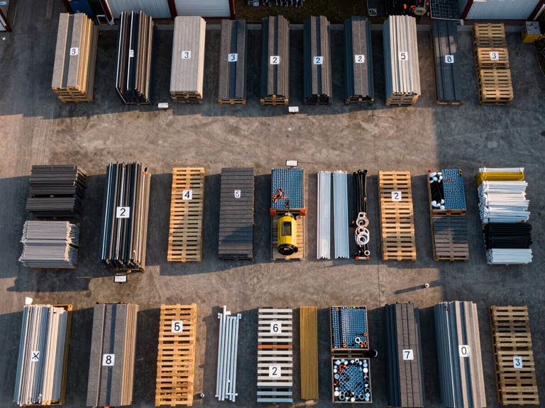 Bird's-eye overhead view of an organized job-site material staging area, pallets sorted by trade — roofing sheets, plumbing fittings, electrical conduit — each labeled with phase markers, golden-hour exterior light casting long shadows across the organized grid