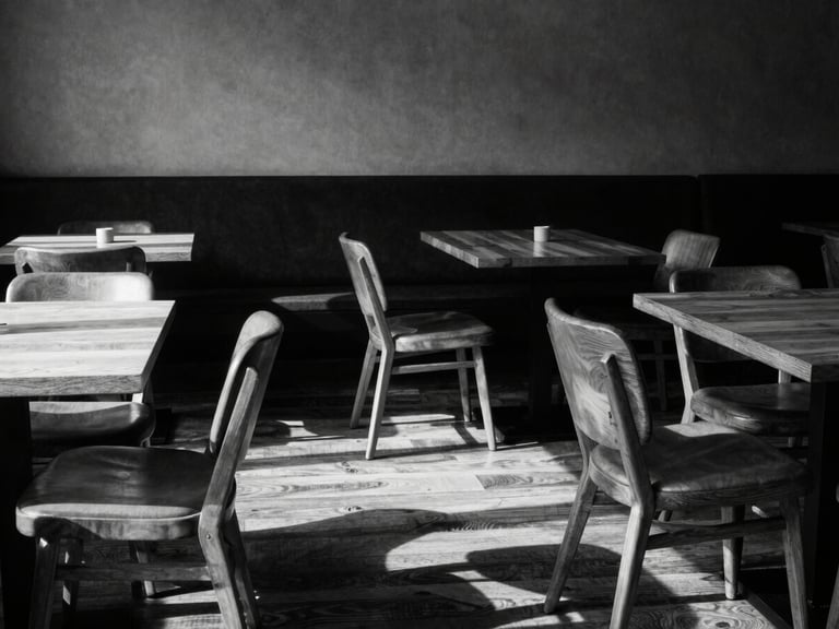 Wide-angle shot of a minimalist café seating area, empty chairs, warm wood tables, strong shadows, high-contrast monochrome tones, 35mm lens.