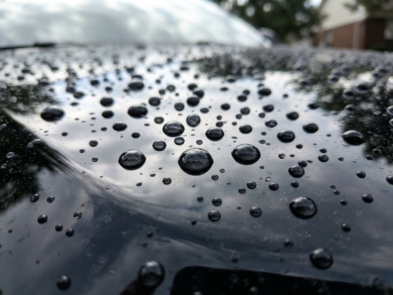 Extreme close-up of water beading on a freshly waxed black SUV hood, high-contrast reflections, bright natural daylight, low angle, sharp focus