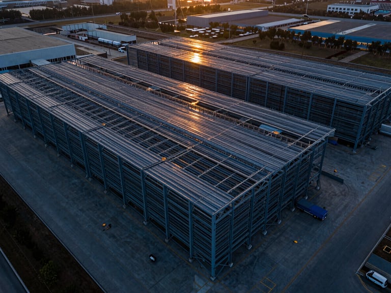 Aerial drone wide-angle shot of a massive industrial logistics park at dusk, steel frame structure visible, dramatic high-contrast lighting with orange sunset reflections on metallic surfaces.
