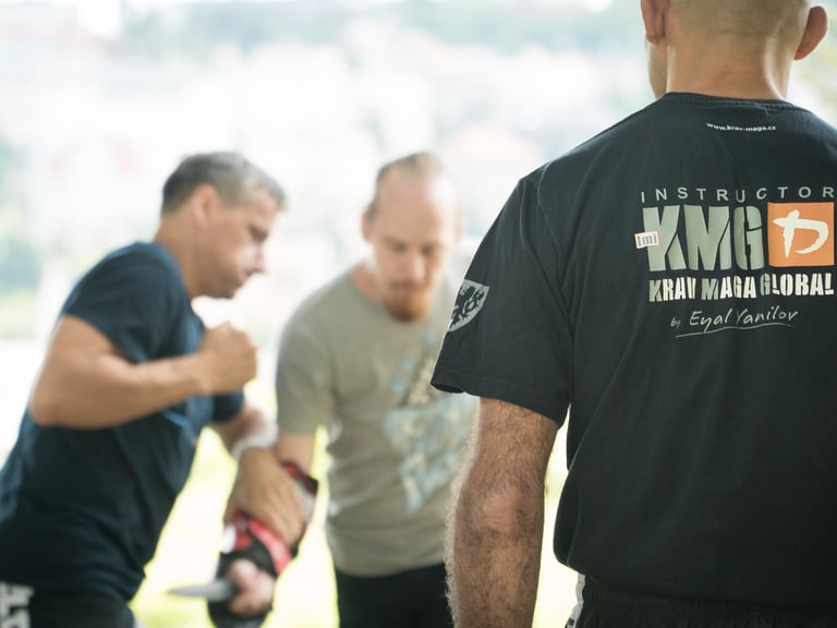 An instructor in a 'Krav Maga Global' shirt observes students practicing self-defense techniques outdoors with a knife.