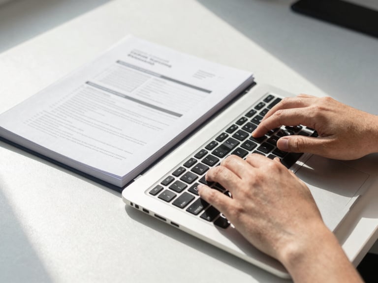 Close-up overhead shot of hands typing on a laptop keyboard in a bright office environment, a printed training workbook open beside the laptop, natural daylight from a nearby window casting clean even light across the desk surface, no faces in frame