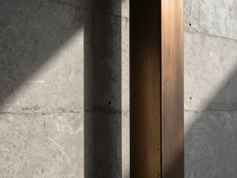 Abstract close-up of a raw concrete wall meeting a brushed bronze structural column, soft natural light casting long linear shadows.