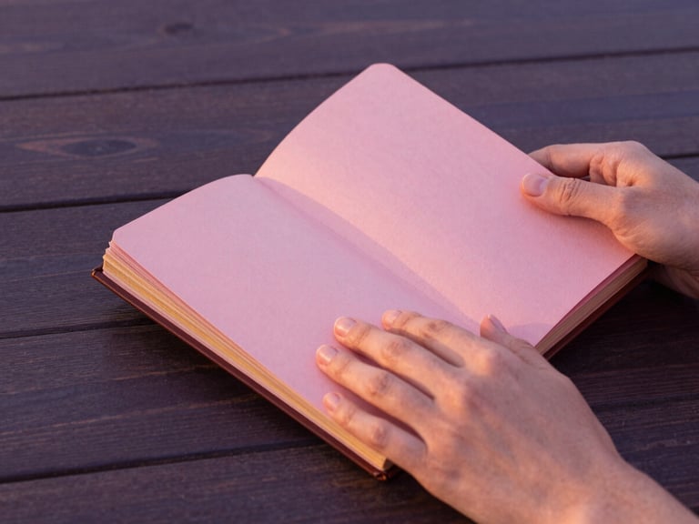 Manos sosteniendo un diario antiguo de cuero, con sutiles detalles en pan de oro en las páginas, sobre una mesa de madera oscura, iluminado por luz suave de atardecer. Primer plano de las manos y el diario, con enfoque asimétrico. Warm editorial photography, soft golden-hour light, dusty pink and deep purple tones, elegant minimalist composition, 35mm.