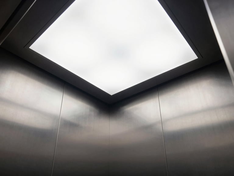 Low-angle interior shot of a premium elevator cabin, brushed steel panels, minimalist LED ceiling grid, sharp reflections, clean architectural lines
