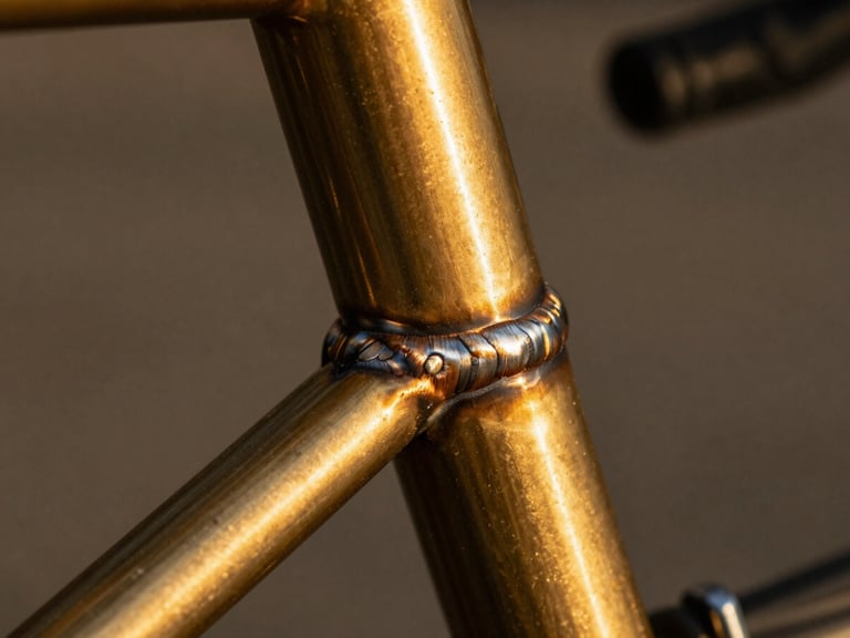 Extreme close-up of a polished brass fillet weld on a bicycle frame, deep shadows, warm editorial photography, 35mm