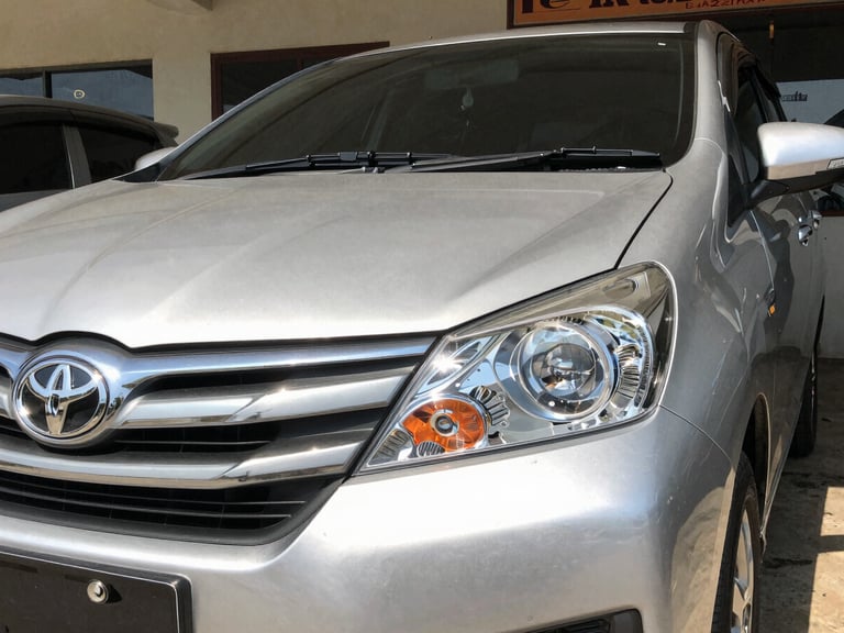 Close up of a clean silver Toyota Avanza front grille and sparkling clean headlight, parked outside a clean local showroom under direct bright Kupang sun, sharp focus