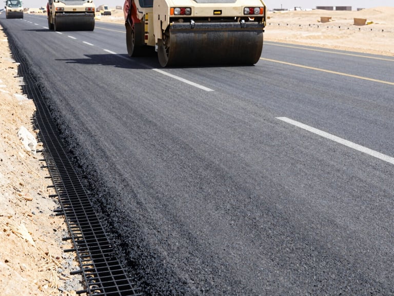 Low-angle wide shot of a multi-lane highway under construction in the Egyptian desert, black fiberglass geogrid mesh visible under a layer of fresh asphalt, heavy road rollers compacting, bright mid-day sun, industrial construction photography.