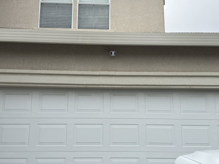 A white sectional garage door under a two-story tan stucco house with a modern security camera installed.