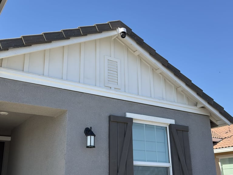 An outdoor security camera mounted on the white gable of a modern grey stucco house for home surveillance.