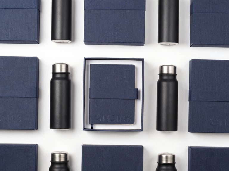 Clean overhead flat-lay of premium linen-wrapped gift boxes, open to reveal debossed journals and matte black thermal flasks, soft studio lighting