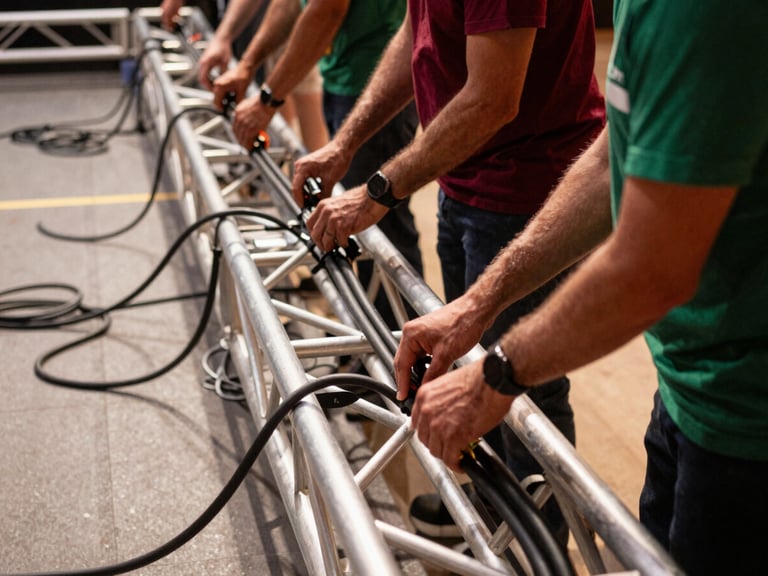 Behind-the-scenes photo of an event crew neatly routing thick black power cables along a metal stage truss, clean workspace, warm natural indoor light, sharp focus, 35mm