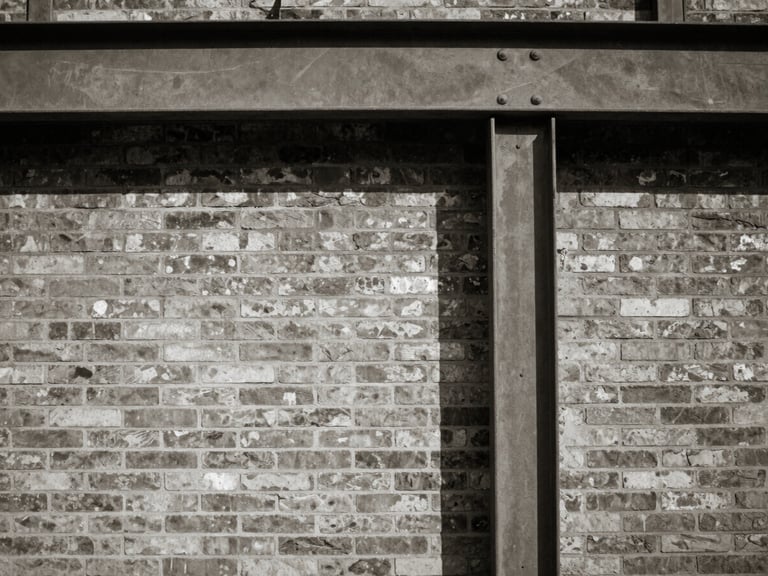 Medium-format photograph of a brick wall meeting a steel beam, high-contrast shadows, raw textures, muted tones.