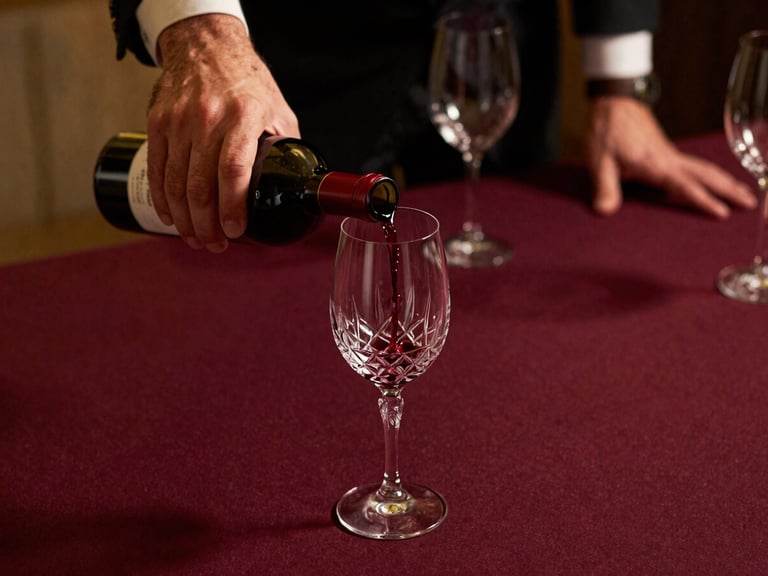 Close-up of a sommelier's hands carefully pouring red wine into a crystal glass at a banquet table, rich maroon tablecloth, low-key golden light, shallow depth of field, 35mm