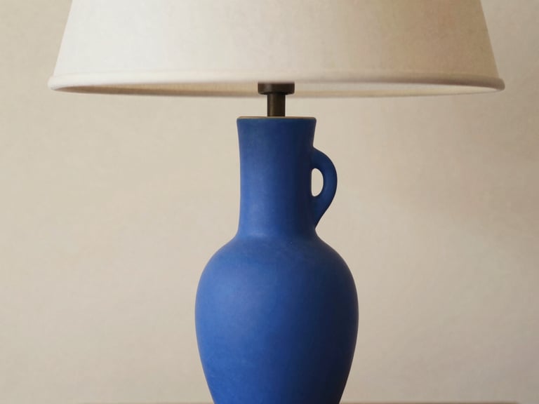 Close-up portrait photograph of a unique, sculptural ceramic table lamp in vibrant blue, minimalist composition on a textured beige background, soft diffused daylight, shallow depth of field, 35mm.