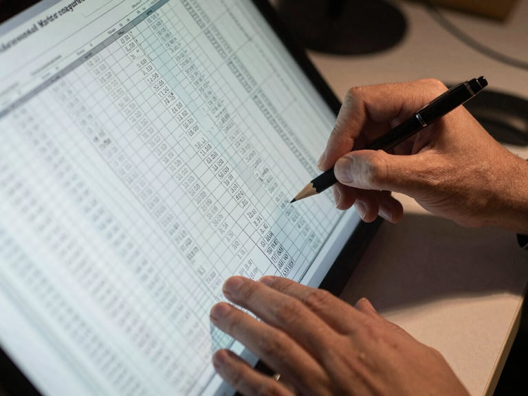 Close-up overhead view of hands annotating a dense financial spreadsheet on a large monitor, a mechanical pencil resting beside printed data rows, cool desk-lamp task lighting casting sharp shadows, paper edge visible at frame bottom