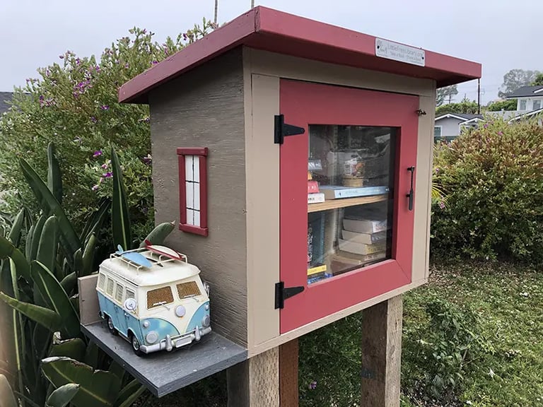A red and tan Little Free Library book sharing box with a miniature blue VW surf van on a side shelf.