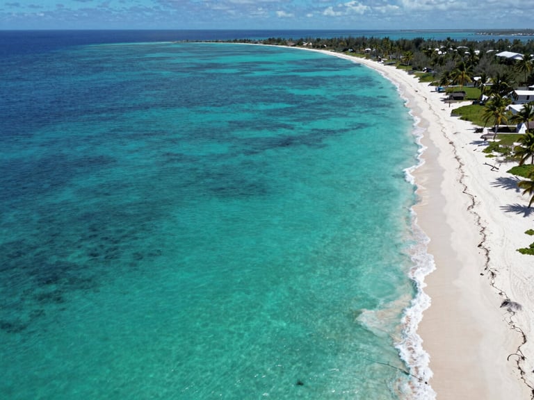 Aerial panoramic view of San Andrés island coastline at midday, vivid turquoise-to-deep-blue gradient sea, a narrow strip of white sand beach visible on the right third, small palm-fringed cays in the distance