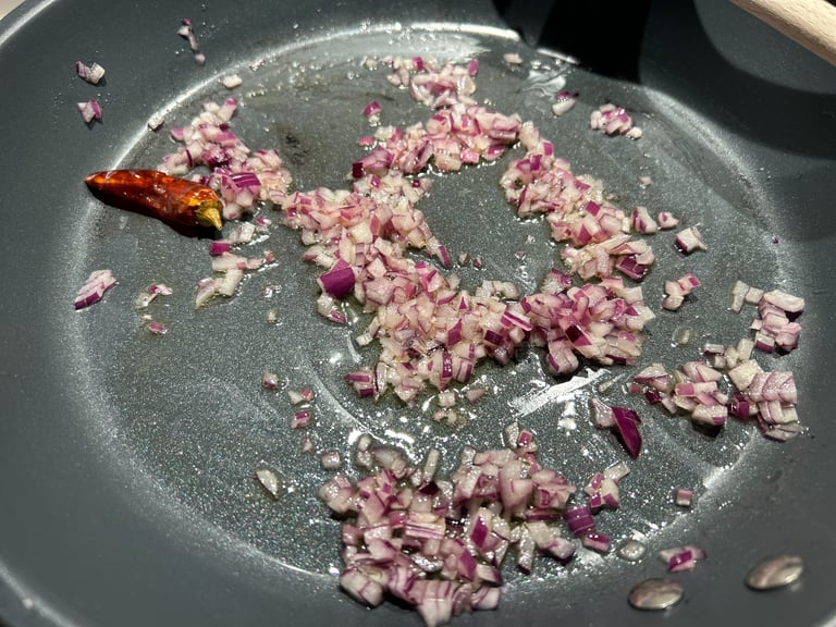 Finely diced red onions and a whole dried chili pepper sautéing in oil in a non-stick frying pan.