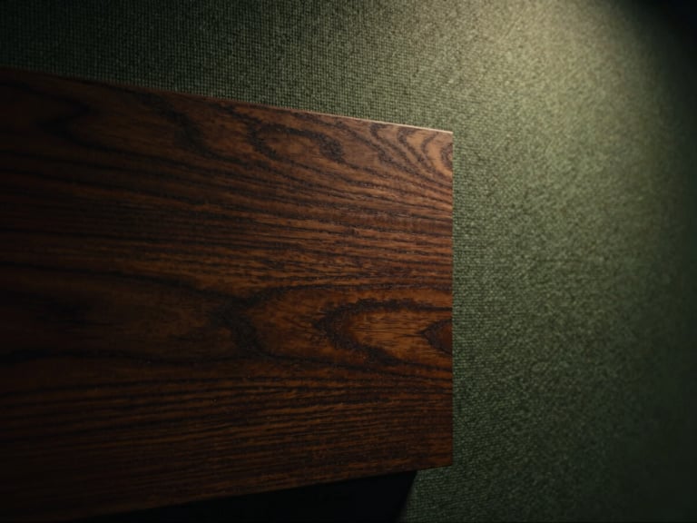 Macro close-up of dark stained walnut wood grain meeting textured acoustic fabric, warm dramatic side lighting, high contrast chiaroscuro, cinematic 35mm rendering