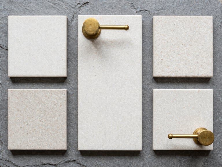 Top-down flat lay of tactile terrazzo tile samples and unlacquered brass fixtures on a neutral slate grey background, bright diffused daylight, sharp focus.