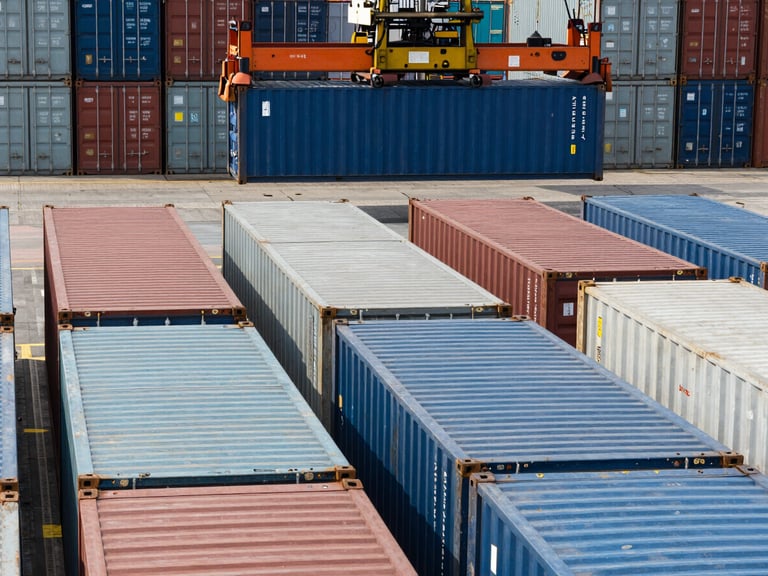 High-contrast daylight shot of neat rows of shipping containers at Port Klang, heavy industrial reach stacker moving a blue container in the background, sharp metallic textures.