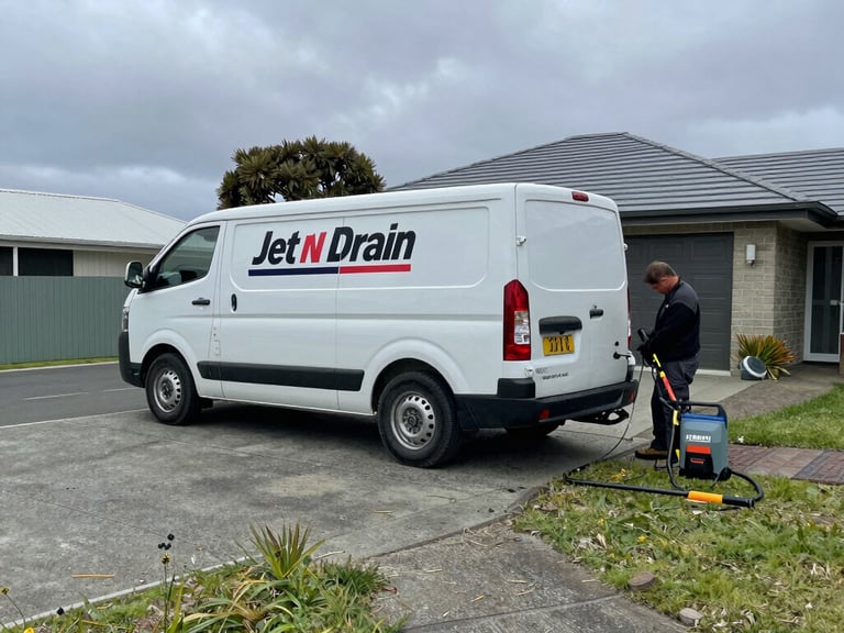 Wide shot of Jet n Drain van parked on a residential driveway, technician working at the rear with jetting equipment laid out, overcast Taranaki daylight, practical documentary framing