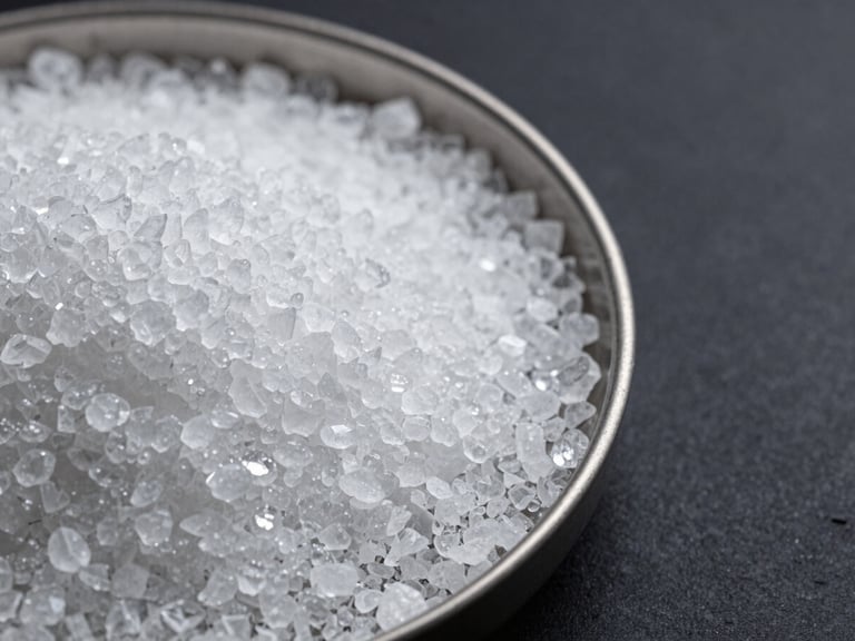 Tight close-up of crystalline lithium hydroxide granules in a shallow steel tray under studio strobe light, individual crystals sharp and bright against a dark industrial surface, framed to the right with dark space left of center