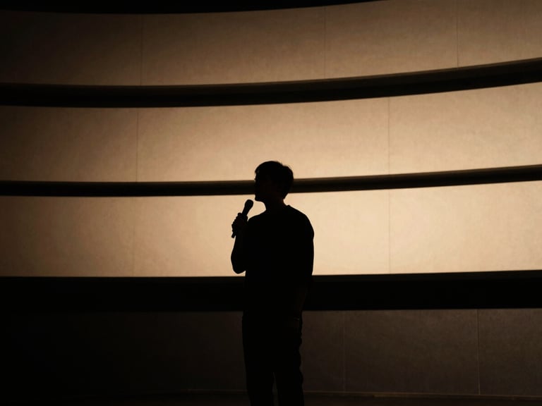 Low-key dramatic portrait of a speaker silhouetted against a warm golden-hour stage light, architectural lines of a modern amphitheater, cinematic atmosphere