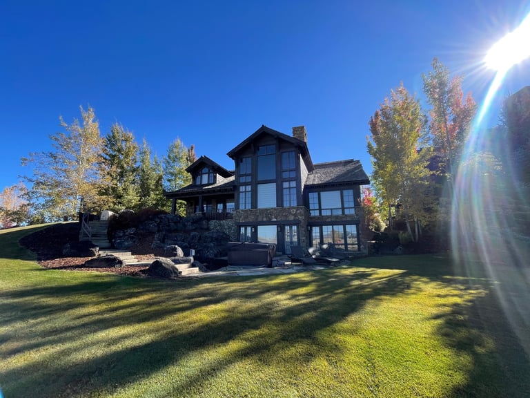 Luxury mountain estate with stone exterior, lush green lawn, and fall foliage under a bright sun.