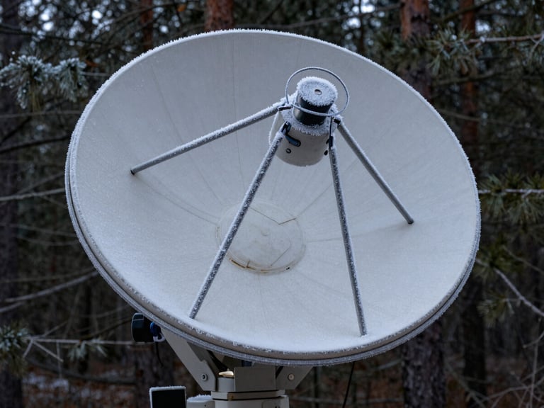 Extreme close-up of a ruggedized satellite dish covered in light frost, mounted on a solid steel bracket against a dark pine forest background.