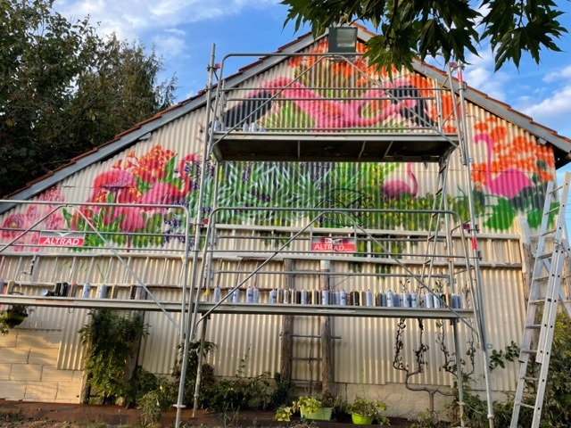 une grange en taule avec un échafaudage et une fresque avec des flamands roses et des plantes
