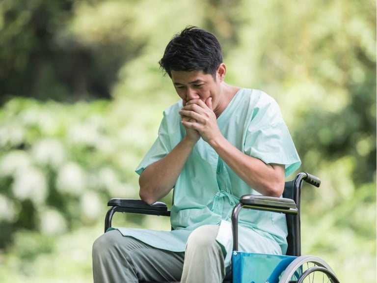 A patient in a hospital gown sitting in a wheelchair outdoors, looking pensive and concerned.