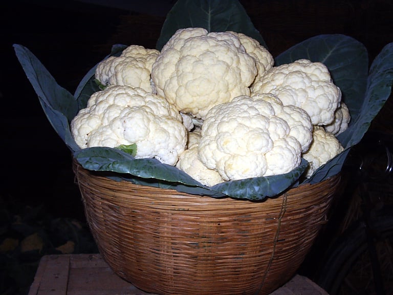 Fresh NXG DELIGHT white cauliflower heads with green leaves in a woven wicker basket at a market.
