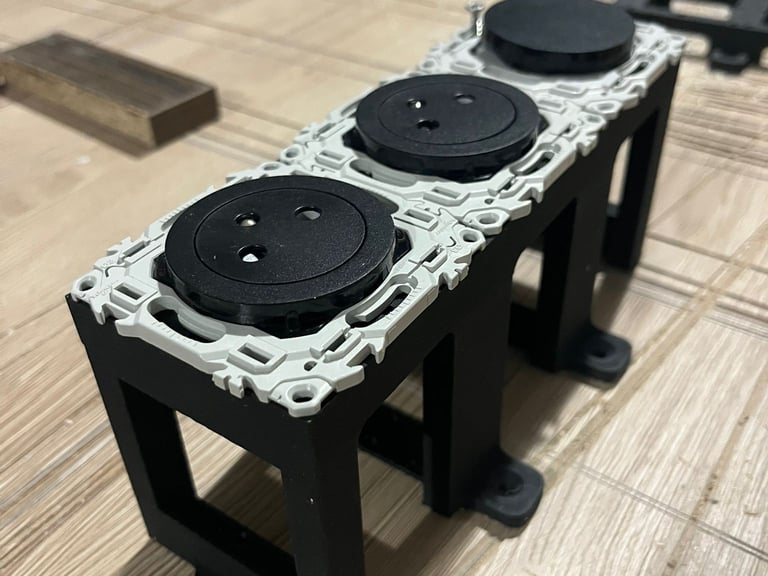 Three-gang black electrical socket outlets mounted on a custom structural bracket on a wooden workbench.