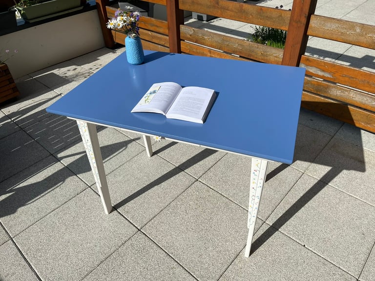 Blue wooden patio table with an open book and a flower vase on a sunny outdoor stone terrace.